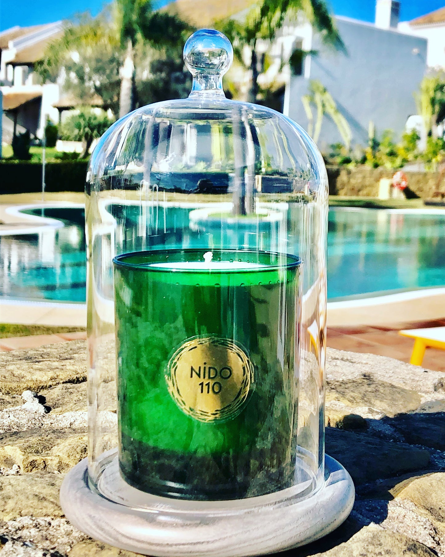Green candle in a glass dome with 'NIDO 110' label by a poolside. dark honey & tobacco scent soy wax