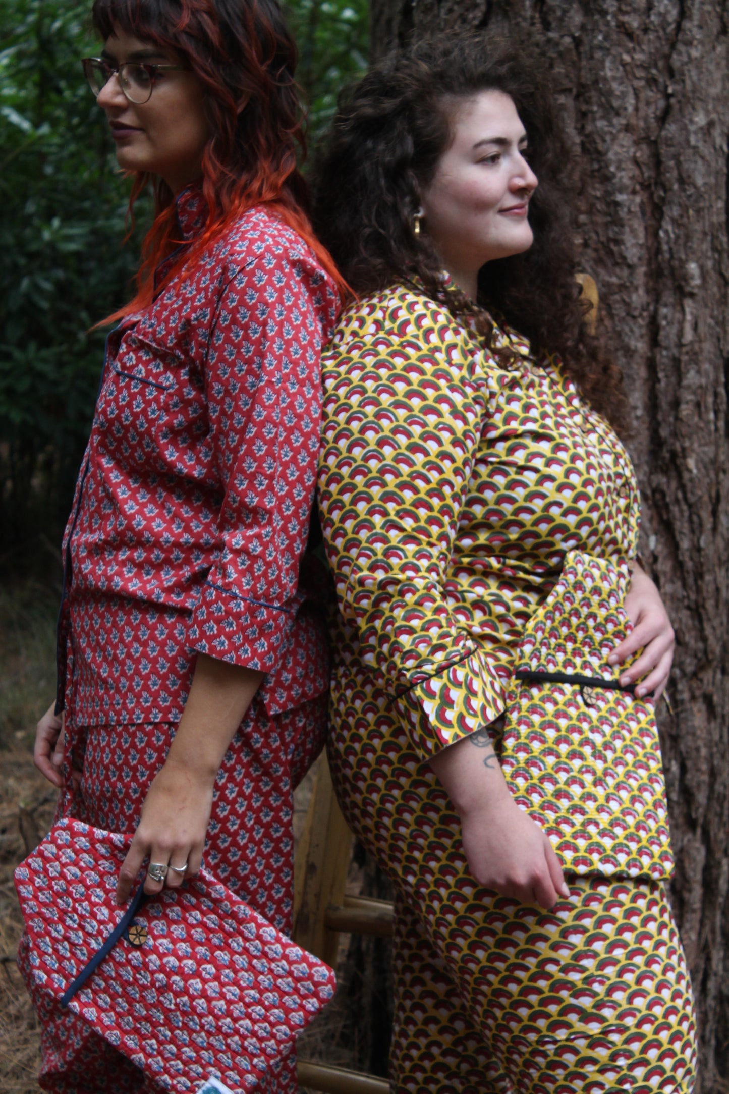 Two models wearing hand block printed pyjamas back to back in different patterns, red, blue, and yellow black and red colours in forest scene holding hot water bottle covers